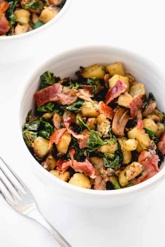 two white bowls with chopped Japanese sweet potatoes, sauteed mushrooms and kale and bacon combined into a breakfast hash. There is a stainless steel fork to the left of the bowl in the center of the image