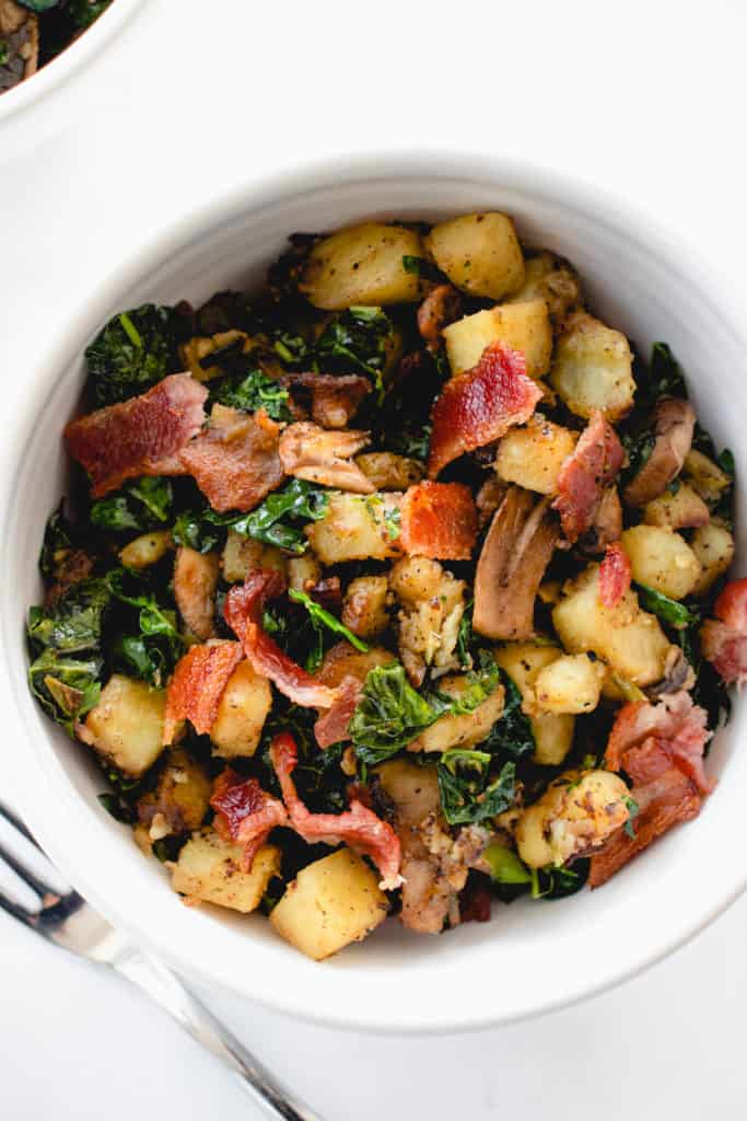 overhead shot of a white bowl with chopped Japanese sweet potatoes, sauteed mushrooms and kale and bacon combined into a breakfast hash. There is a stainless steel fork to the left of the bowl in the center of the image