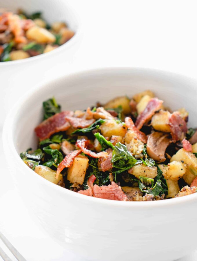 two white bowls with chopped Japanese sweet potatoes, sauteed mushrooms and kale and bacon combined into a breakfast hash. There is a stainless steel fork to the left of the bowl in the center of the image