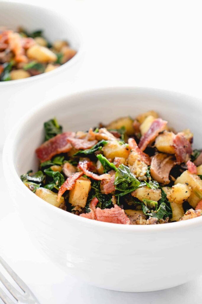 two white bowls with chopped Japanese sweet potatoes, sauteed mushrooms and kale and bacon combined into a breakfast hash. There is a stainless steel fork to the left of the bowl in the center of the image