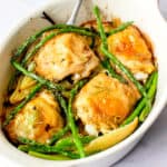 white oval shaped baking dish with four chicken thighs, asparagus and fennel with a stainless steel serving spoon against a white and grey marbled background