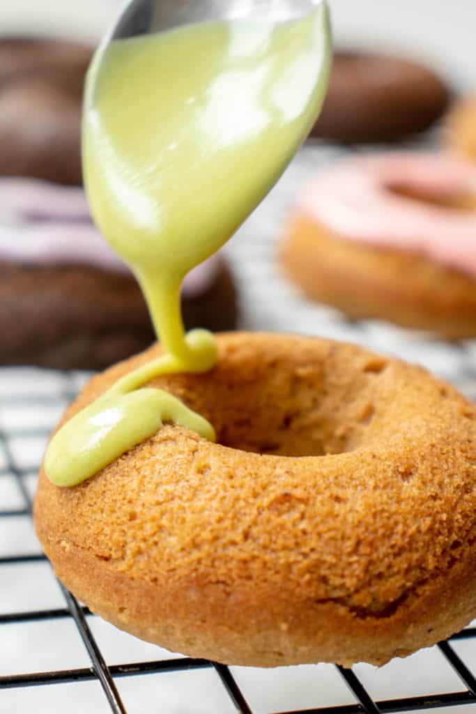 black cooling rack with pastel-glazed mini donuts in the background and a vanilla donut being drizzled with a bright green icing with a spoon in the foreground