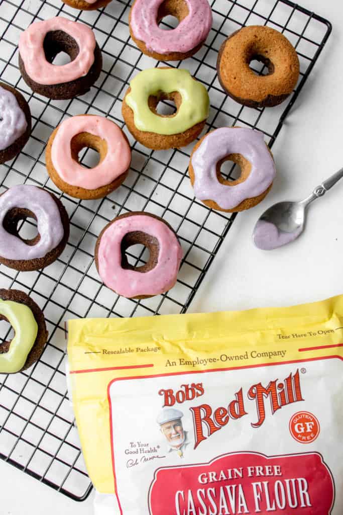 a variety of pastel colored mini donuts sitting on top a wooden cutting board lined with a white piece of parchment paper next to a black cooling rack with some more donuts and a yellow, red and white bag of bob's red mill cassava flour on the bottom right corner of the frame
