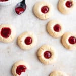 overhead shot of paleo thumbprint cookies filled with berry filling on a light beige textured background. there is a bite taken out of one of the cookies, a small black spoon with a bit of berry filling and a small white bowl filled with berry filling in the top left corner. in the top right corner, a black wire cooling rack is visible at a diagonal