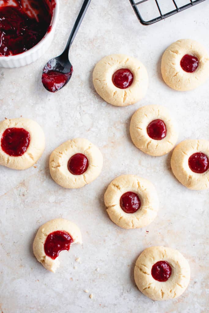 overhead shot of paleo thumbprint cookies filled with berry filling on a light beige textured background. there is a bite taken out of one of the cookies, a small black spoon with a bit of berry filling and a small white bowl filled with berry filling in the top left corner. in the top right corner, a black wire cooling rack is visible at a diagonal