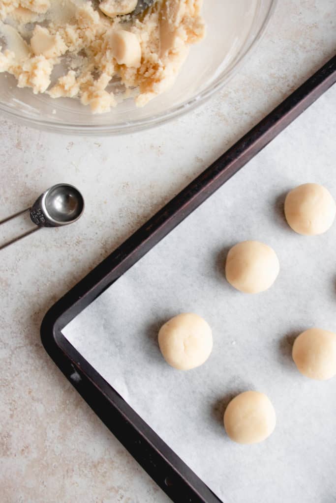overhead shot of a sheet pan lined with white parchment paper with balls of cookie dough. To the left of the cookie sheet is a stainless steel teaspoon and a clear glass bowl with cookie dough