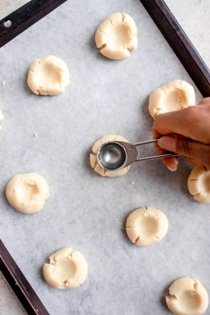 sheet pan placed on a diagonal, lined with white parchment paper and balls of dough with an indentation in the middle. there is a black hand pressing down a teaspoon in the middle of one cookie to make an indentation