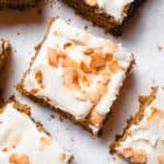 overhead shot of squares of paleo carrot cake topped with buttercream frosting and toasted coconut flakes on a white piece of parchment paper