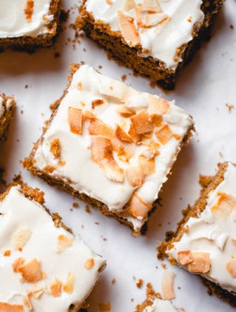 overhead shot of squares of paleo carrot cake topped with buttercream frosting and toasted coconut flakes on a white piece of parchment paper