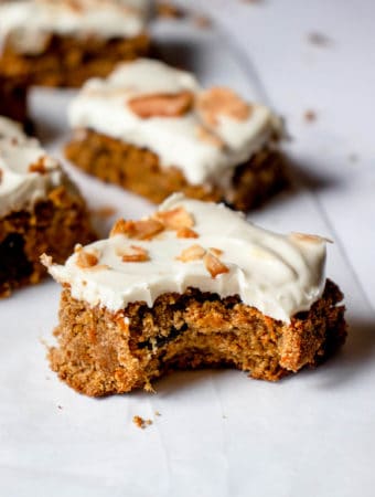 squares of paleo carrot sheet cake scattered on a piece of parchment paper. the square closest to the front of the image has a bite taken out of it revealing its texture