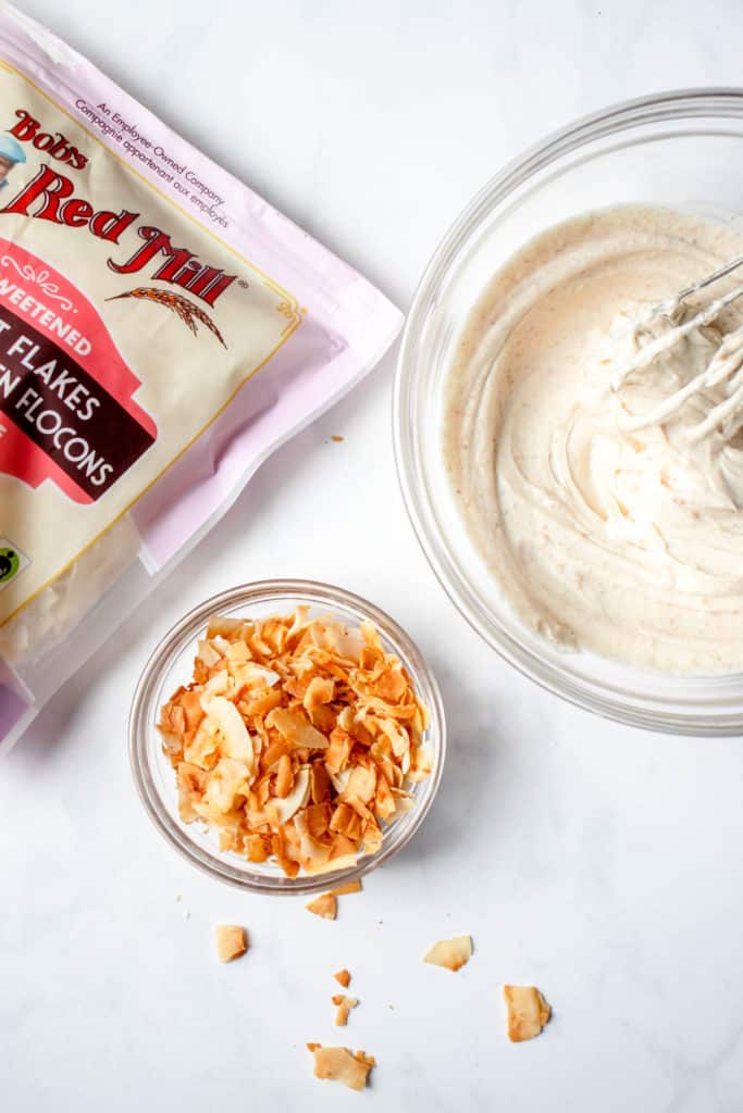 overhead shot of clear glass bowl with toasted coconut flakes. There are some scattered toasted flakes around the bowl. To the left of the bowl is a purple, red, pink, white and brown Bob's Red Mills bag of unsweetened coconut flakes and to the right is a bowl of buttercream frosting