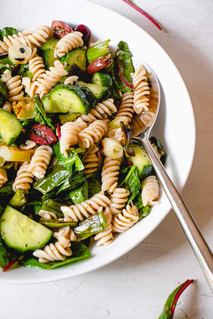 close up shot of a white plate with vegan pasta salad consisting of grain-free pasta, olives, cucumbers, spinach, pickled red onions and grilled green onions.