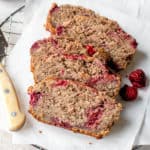 overhead shot of five slices of lemon raspberry loaf lying on two sheets of white parchment paper on a circular metal cooling rack. There is a bone-colored knife to the left of the slices of bread and two raspberries to the right of the slices. In the background of the photo on the left hand side is a white textured bowl with raspberries