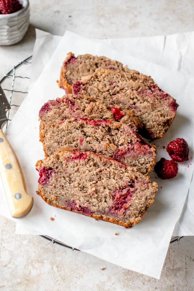 overhead shot of five slices of lemon raspberry loaf lying on two sheets of white parchment paper on a circular metal cooling rack. There is a bone-colored knife to the left of the slices of bread and two raspberries to the right of the slices. In the background of the photo on the left hand side is a white textured bowl with raspberries