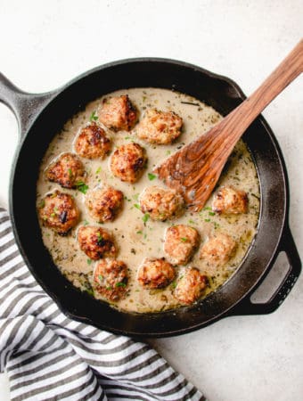 black cast iron skillet with chicken meatballs in cream mushroom sauce with wooden spoon on a grey background with a grey and white cloth napkin on the bottom left