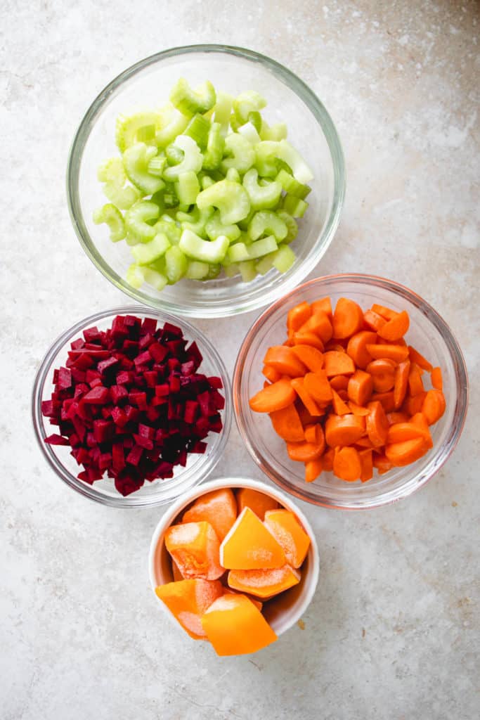 overhead shot of three glass bowls filled with chopped celery, carrots and beets and a small white ceramic bowl with chopped frozen butternut squash. the bowls are tightly framed vertically against a light beige background