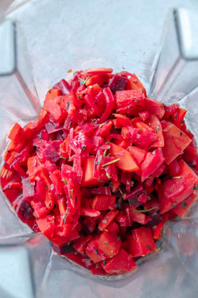 overhead shot of blender with cooked beets, carrots, celery, onions, garlic, butternut squash and herbs which have all taken on a red/purple hue from the beets