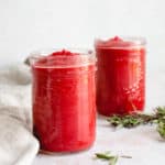 two mason jars filled with nomato sauce diagonally placed on a beige table top with a light beige cloth napkin to the left of the jars there are two sprigs of fresh rosemary placed to the right or the jars and a few leaves of rosemary scattered toward the front of the frame