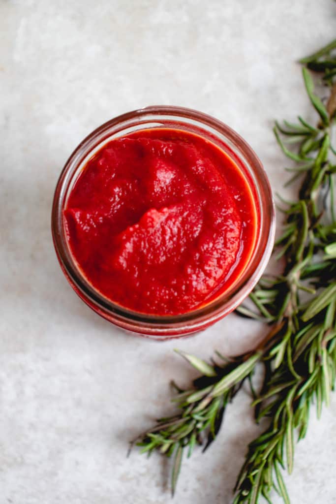 overhead shot of a mason jar with nomato sauce on a light beige background with two sprigs of rosemary to the right of the jar