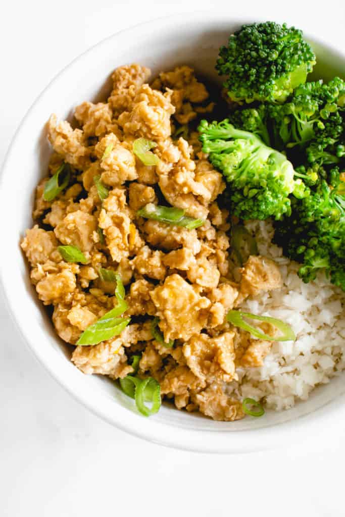 white bowl with ground chicken in orange sauce topped with sliced green onions, steamed broccoli and cauliflower rice against a white background