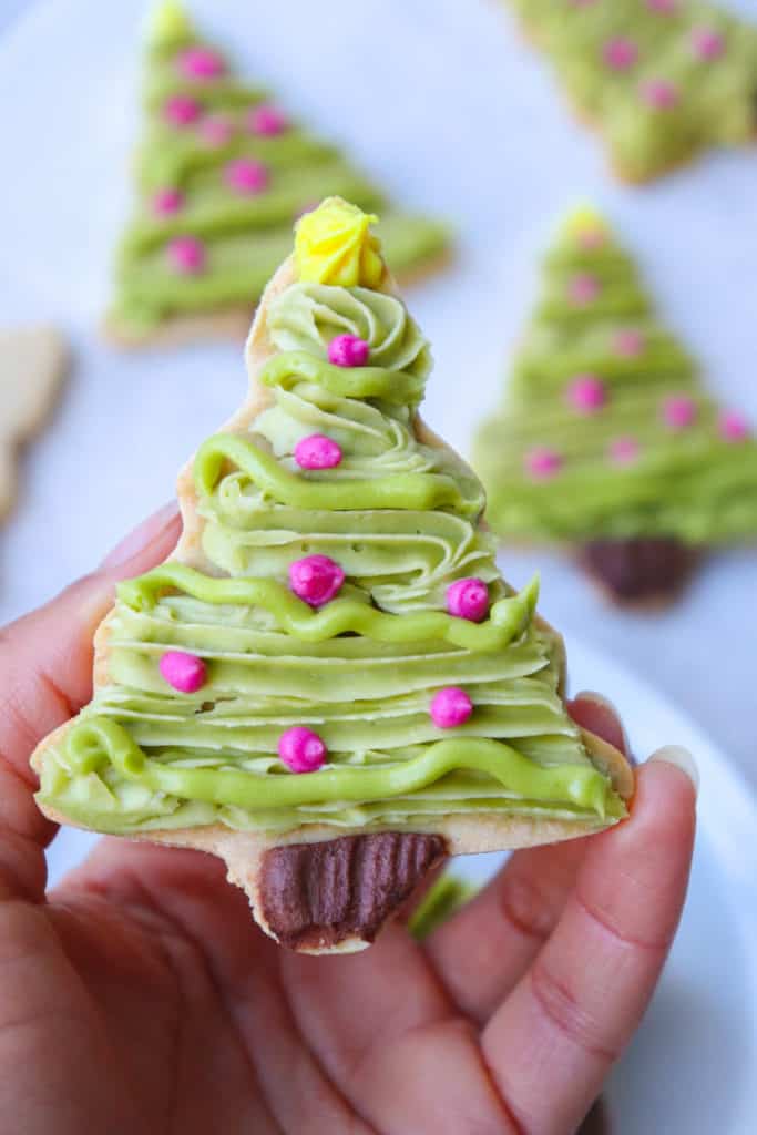 hand holding christmas tree cookie decorated with green, brown, pink and yellow icing with more cookies in the background, blurred