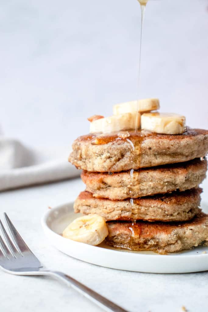 white plate with stack of four banana pancakes topped with sliced bananas, with a small fork to the left and a beige cloth napkin in the background with a drizzle of maple syrup
