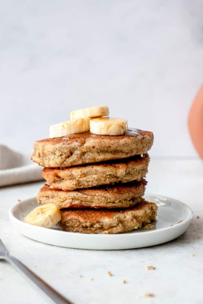 white plate with stack of four banana pancakes topped with sliced bananas, with a small fork to the left and a beige cloth napkin in the background and the side of a terracotta vase