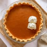 pumpkin pie with two dollops of coconut whipping cream in glass pie dish on top burlap place mat with stainless steel pie knife, a beige cloth napkin and three cinnamon sticks in the lower half of the image, a small gourd on the top left and white plate on the top right