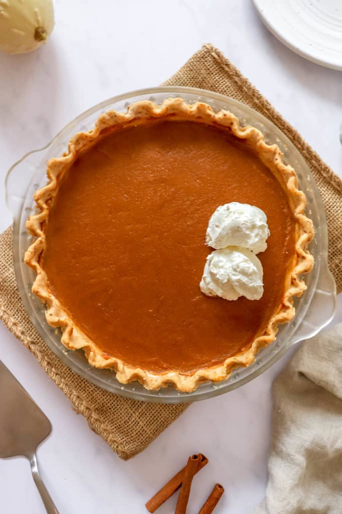 pumpkin pie with two dollops of coconut whipping cream in glass pie dish on top burlap place mat with stainless steel pie knife, a beige cloth napkin and three cinnamon sticks in the lower half of the image, a small gourd on the top left and white plate on the top right