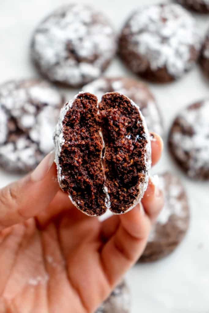 hand holding a gluten-free, AIP/paleo chocolate crinkle cookie broken in half to reveal the fluffy interior texture above a baking tray of crinkle cookies