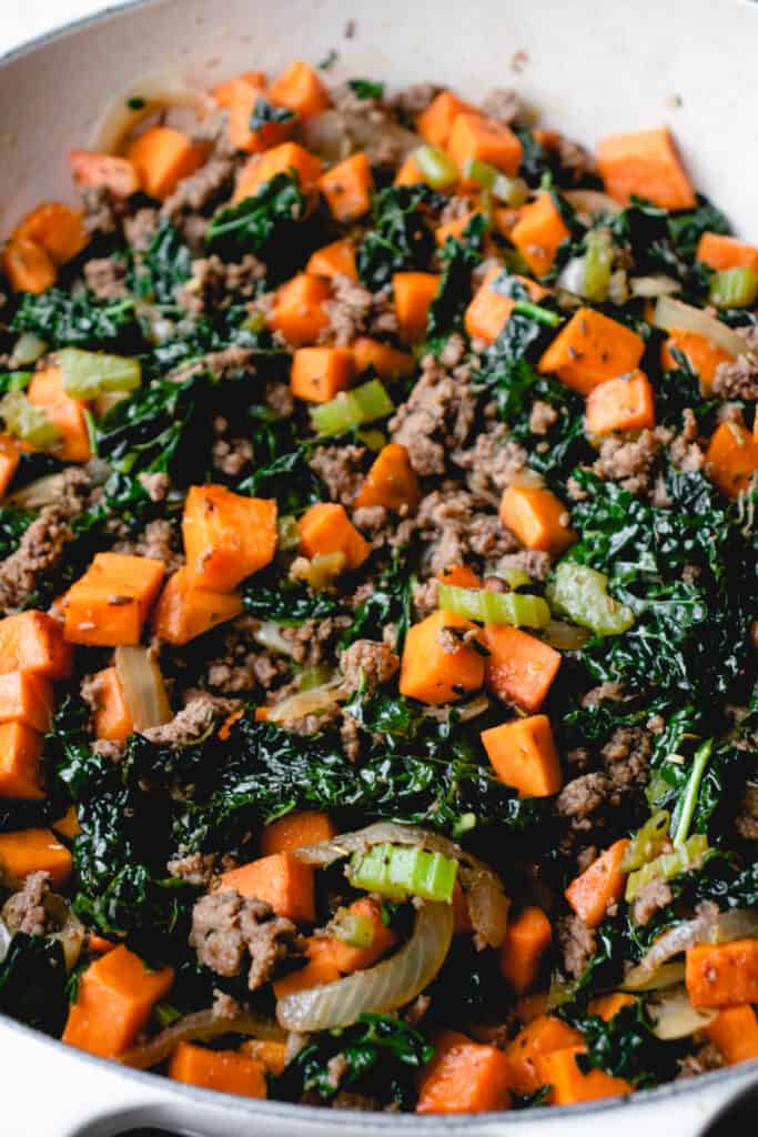 chopped orange sweet potatoes, kale and ground beef sautéing in a cream enamel coated cast iron pot with a wooden spoon
