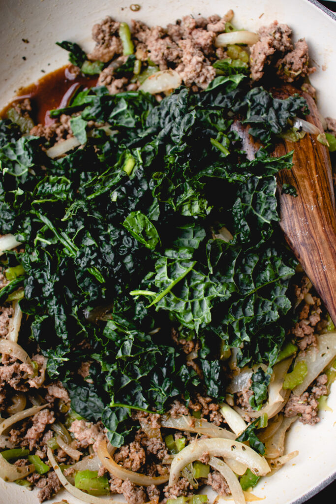 kale, celery, sliced onions and ground beef in a cream enamel coated in cast iron pot with a wooden spoon
