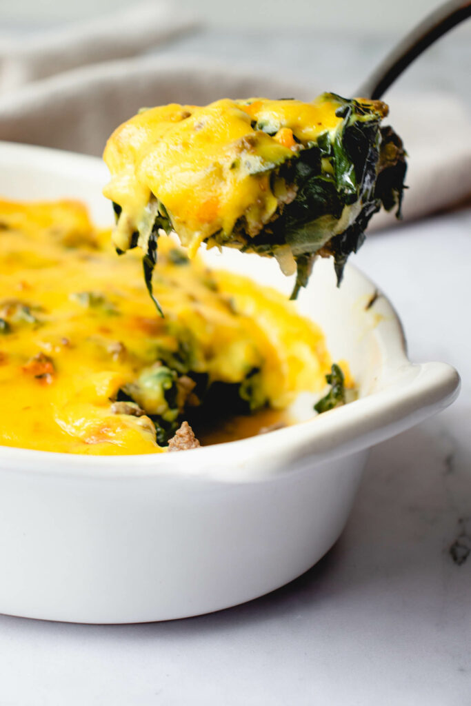 oval cream casserole dish with beef and kale topped with dairy free cheese sauce. a serving spoon is shown lifting a portion of the casserole out the dish