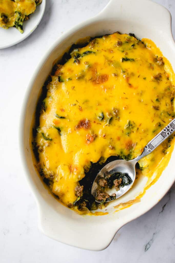 overhead shot of oval shaped cream casserole dish with dairy free cheese topped casserole dish. One portion is scooped out to reveal the ground beef and kale inside and there is a stainless steel spoon resting in the dish
