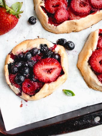 three mini galettes with strawberries and blueberries with a few blueberries and a strawberry scattered around the galettes on a white piece of parchment paper inside a brown baking sheet