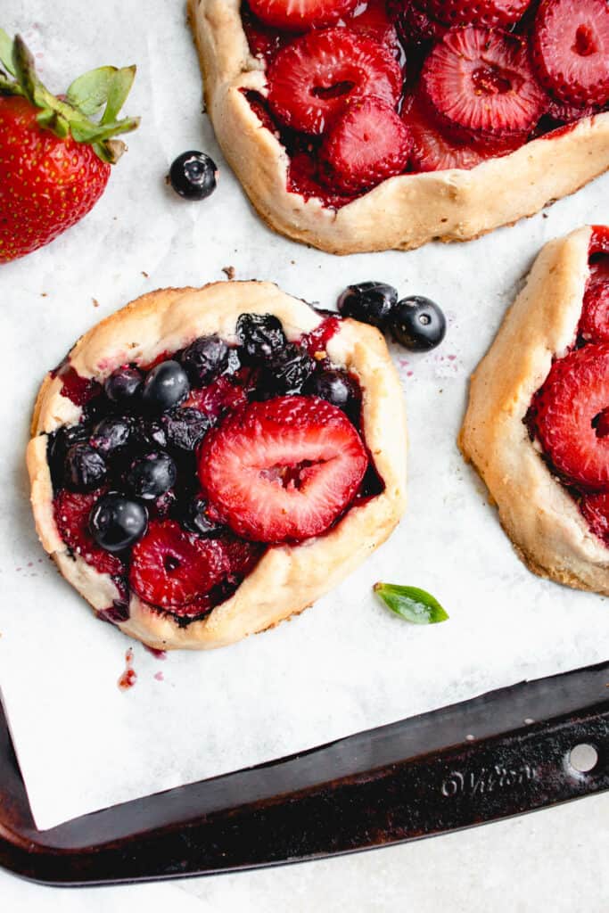 three mini gluten-free galettes with strawberries and blueberries with a few blueberries and a strawberry scattered around the galettes on a white piece of parchment paper inside a brown baking sheet