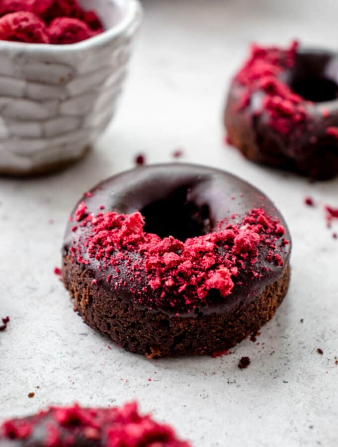 three baked chocolate donuts with raspberries on a light grey background with a white bowl of freeze dried raspberries in the background