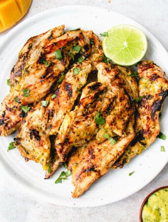 White plate with strips of bbq chicken breasts seasoned in allergy-free mango lime sauce, garnished with a chopped cilantro and a lime wedge. the plate is surrounded by a small wooden bowl with marinade and a half a mango