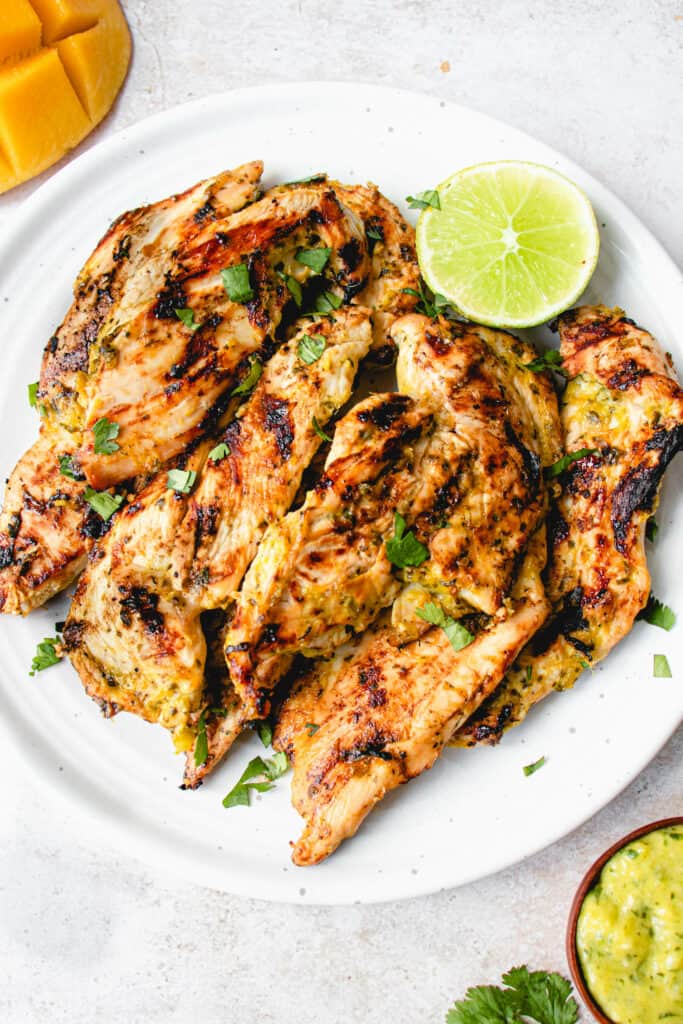 White plate with strips of bbq chicken breasts seasoned in allergy-free mango lime sauce, garnished with a chopped cilantro and a lime wedge. the plate is surrounded by a small wooden bowl with marinade and a half a mango