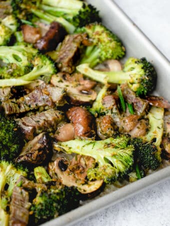 stainless steel sheet pan with beef, broccoli and mushrooms at an angle against a speckled grey background