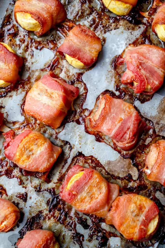 round 1/2 inch slices of plantain wrapped in cooked bacon on a baking sheet lined with white parchment paper. There are caramelized pieces of plantain and burnt bacon fat around the appetizers