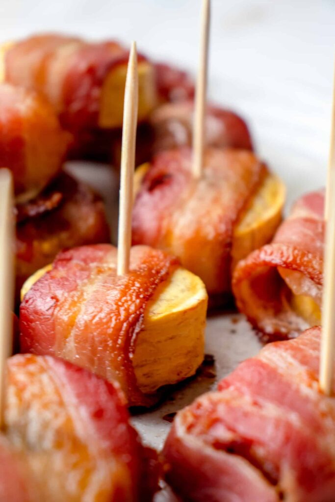 beige speckled plate with slices of sweet baked plantains wrapped in bacon. There are wooden toothpicks placed into some of the appetizers for serving