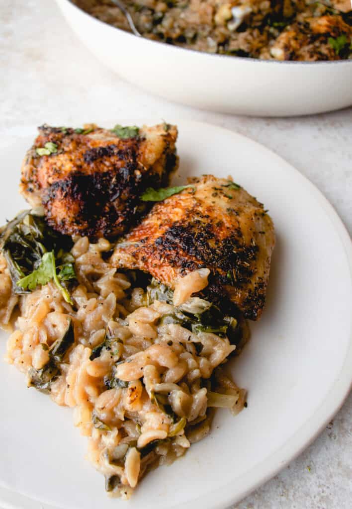 White plate with two chicken thighs, cassava orzo and spinach, garnished with parsley. In the background is a cast iron pan with chicken and cassava orzo dish.