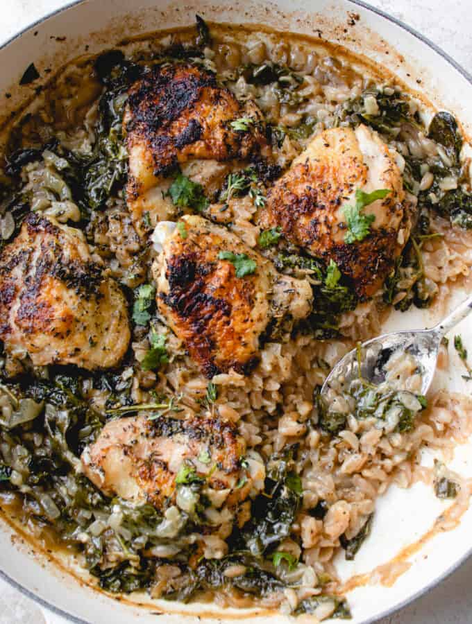 Cast iron pan with baked chicken thighs, cassava orzo and spinach, with a silver spoon laid in the pan.