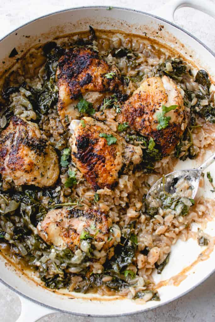 Cast iron pan with baked chicken thighs, cassava orzo and spinach, with a silver spoon laid in the pan.