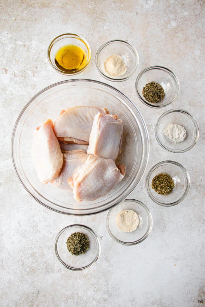 Overhead shot of herb marinade ingredients in small glass bowls, and chicken thighs in a large glass bowl.