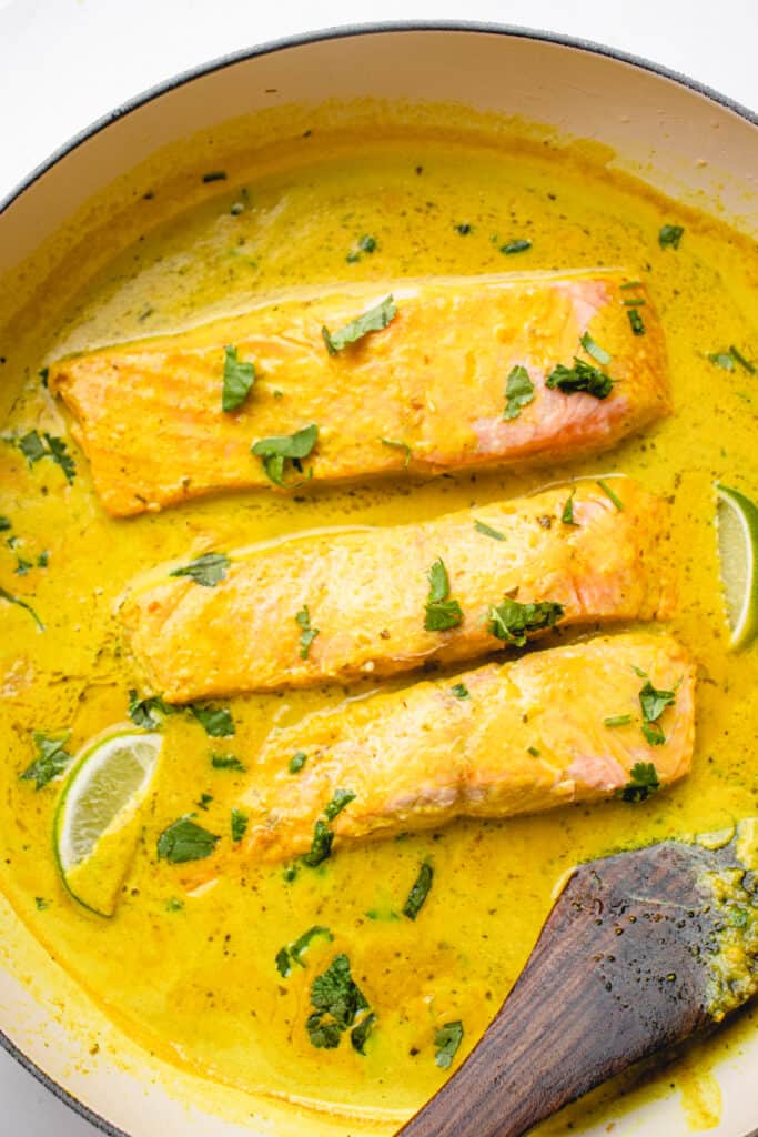 Skillet with salmon filets in coconut curry sauce and garnished with fresh herbs and lime wedges.