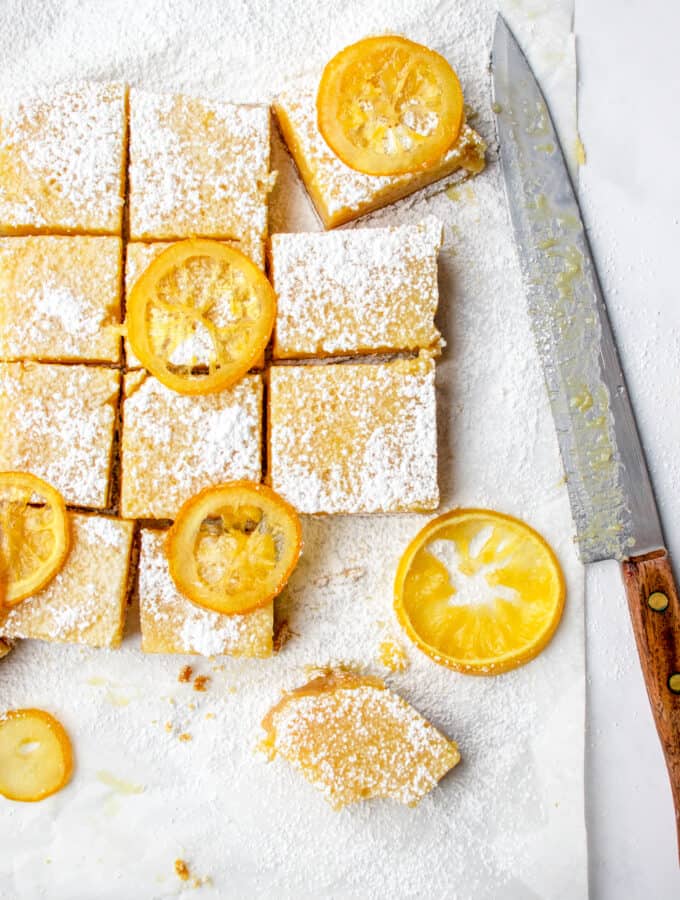 Parchment paper with cut lemon squares, topped with dusted arrowroot starch and baked lemon slices. A large, wooden handled knife is placed on the edge, after cutting.