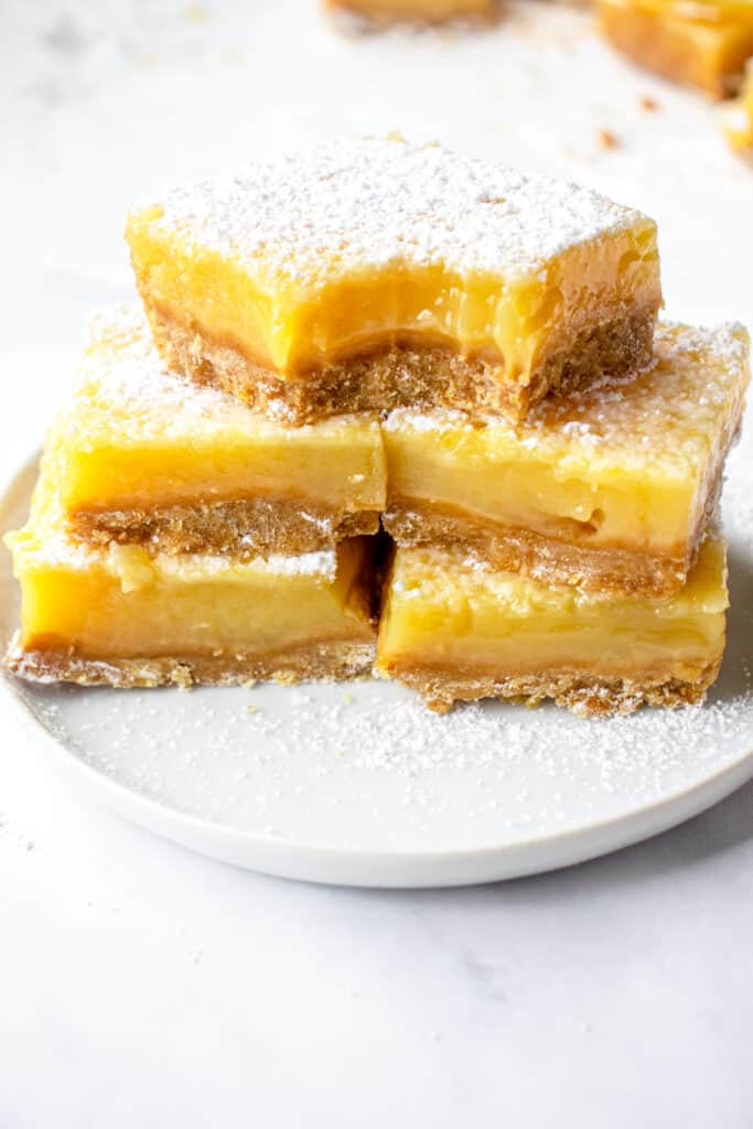 A small white plate with a two stacks of two lemon bars. The fifth lemon bar placed atop the middle of the stacks is bitten into, to show texture.