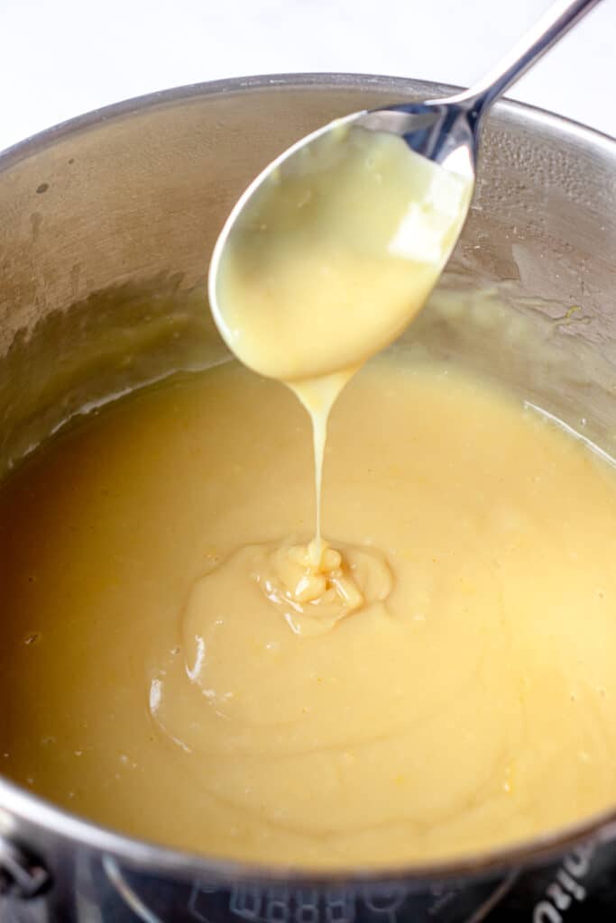 A saucepan with the lemon filling. A metal spoon is coated with the lemon curd and held up out of the pan, to show the lemon filling's thickness.
